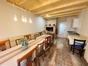 A dining area with a long wooden table, chairs, hanging light fixtures, a fireplace, and a kitchen area with a sink and cabinets. - Chalet 'Casa Rural La Abuela' with Mountain Views, Wi-Fi, and Air Conditioning (Mainar)