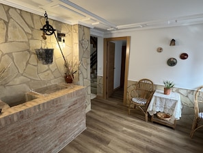 Un comptoir d’accueil en pierre, surmonté d’un luminaire suspendu, une chaise en osier, une petite table avec une plante, et un escalier en arrière-plan.