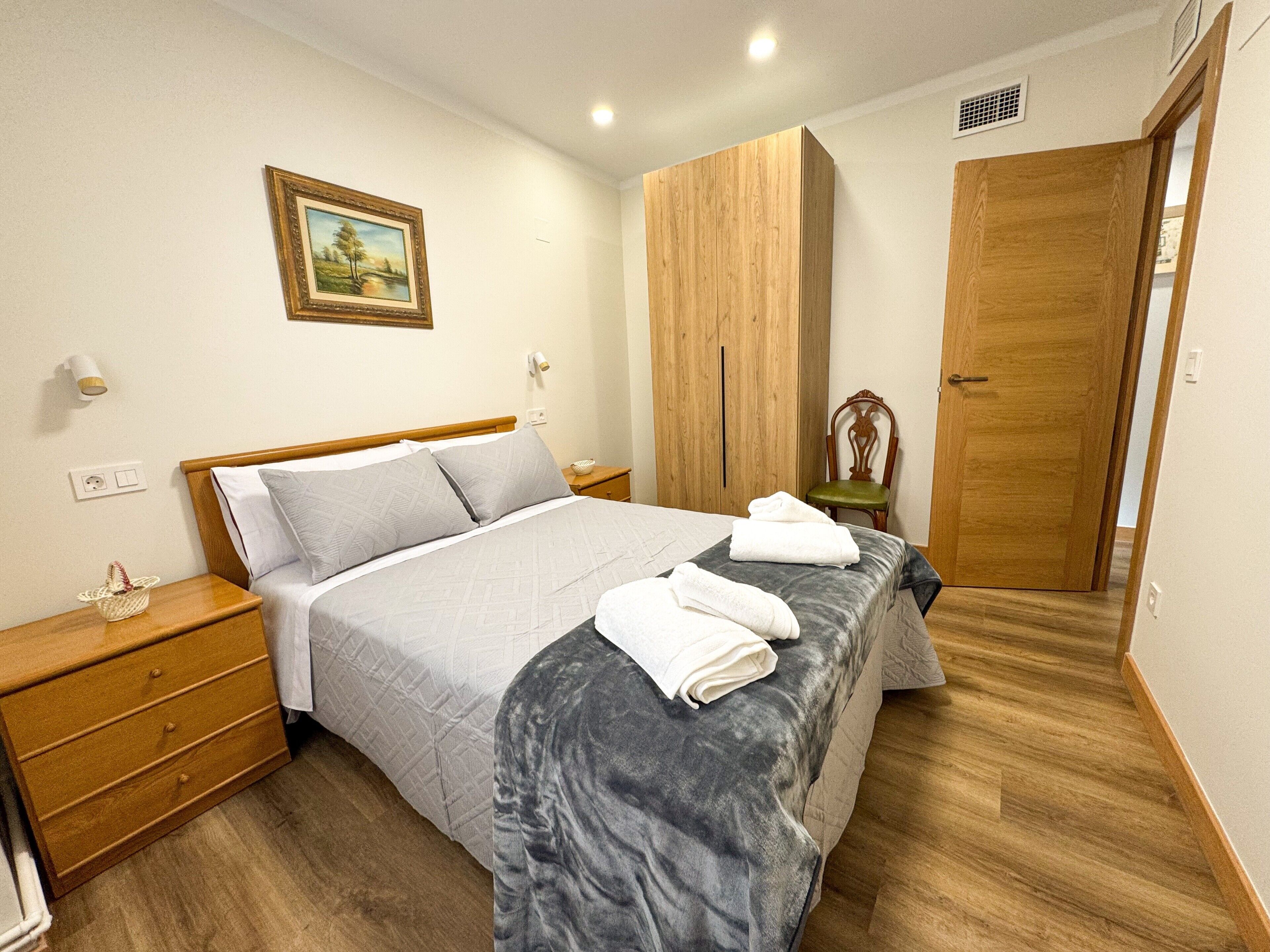 A bedroom with a bed, a nightstand, a wooden wardrobe, a chair, and a door.