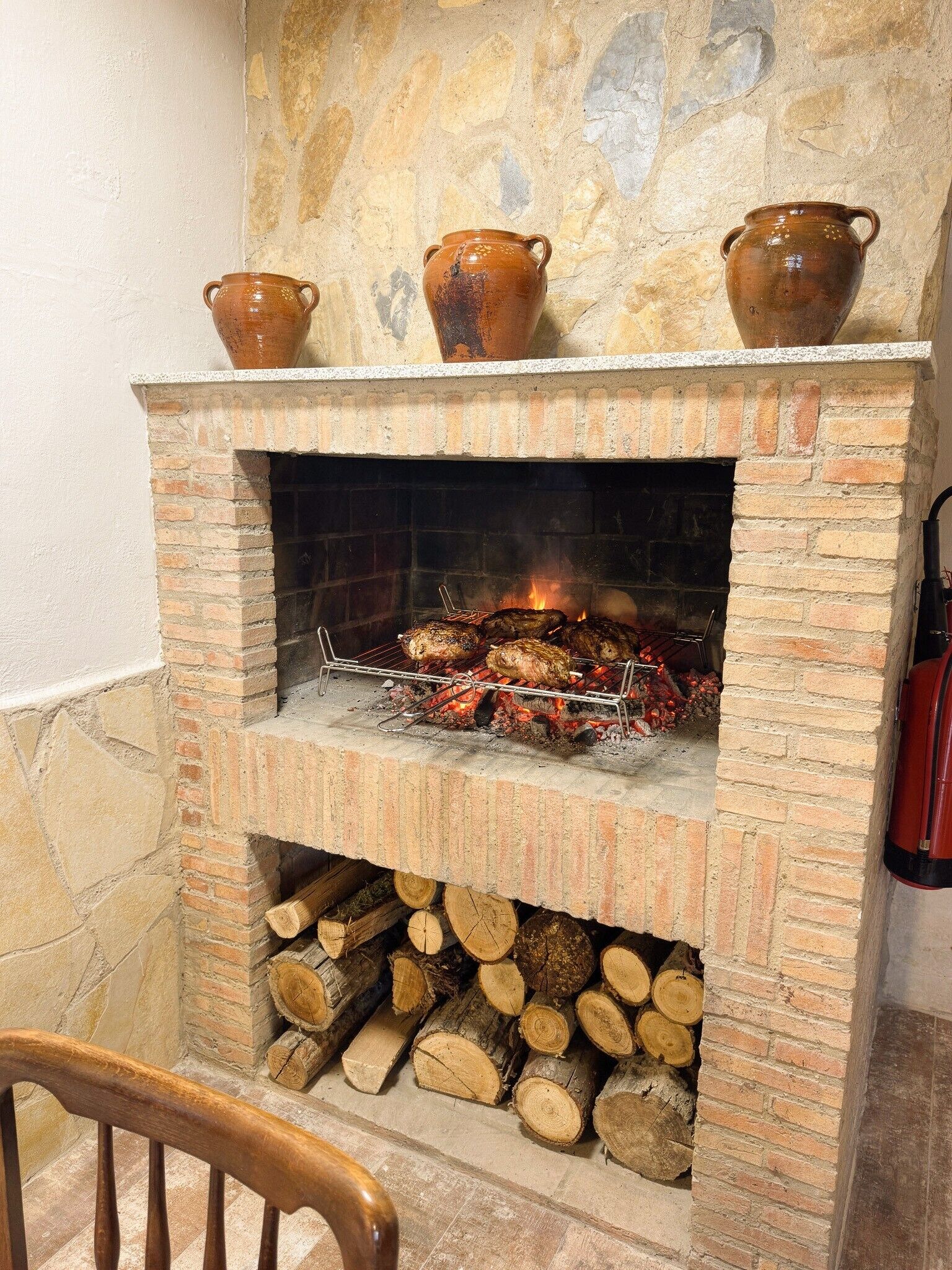 A brick fireplace with a fire, a wooden chair in front, and stacked firewood.