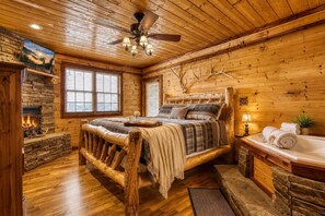 A log cabin bedroom with a stone fireplace, a bed with plaid bedding, a wooden headboard, and a ceiling fan. - Smoky Sunset Retreat, Hot Tub, Mountain Views, Free Tickets! (Sevierville)