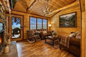 Interior de una cabaña de troncos con chimenea, muebles de madera, un candelabro y una pintura de un paisaje montañoso.