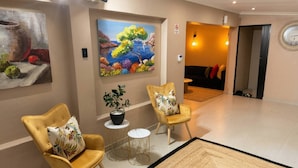 Een moderne hotellobby met twee gele fauteuils, een bijzettafeltje met een plant, een schilderij van een kustlandschap en een klok aan de muur.