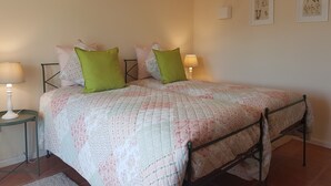 A bedroom with a bed, two bedside tables with lamps, a wall with framed botanical prints, and a tiled floor.