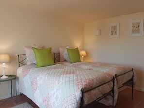 A bedroom with a bed, two bedside tables with lamps, a wall with framed botanical prints, and a tiled floor.