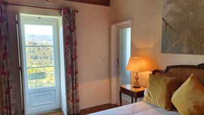 A bedroom with a bed, a nightstand with a lamp, a door, and a view of the outdoors.