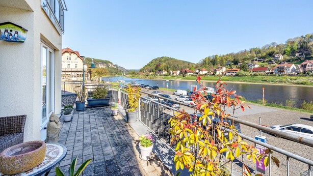 Ein Balkon mit Blumentöpfen, Blick auf einen Fluss und ein Gebäude im Hintergrund, an dem das Schild 'TEL CAFE RICHTER' befestigt ist.