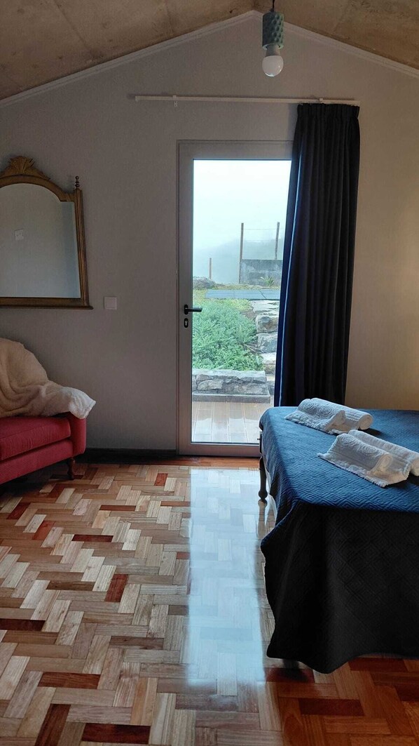 A room with a bed, a red sofa, a mirror, and a view through a glass door to an outdoor area.