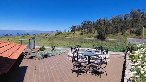 A patio with a table and chairs, a view of the landscape, and a red-roofed building. - Casas do Pargo- Palheiro (Lombada Velha)