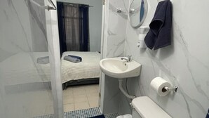 Un baño con ducha, inodoro, lavamanos y una cama visible al fondo.