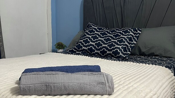 Una cama bien tendida con una colcha blanca y gris, un cabecero gris y almohadas estampadas.