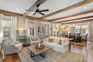 A spacious living room with a high ceiling, wooden beams, and a ceiling fan. There is a dining area with a table and chairs, and a large window providing natural light. - WaterSound Retreat | Private Pool | Near 30A Beach (Watersound)