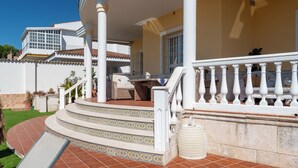 Un espacio al aire libre techado con una escalera que conduce a un balcón, un sofá de mimbre y una pequeña mesa.