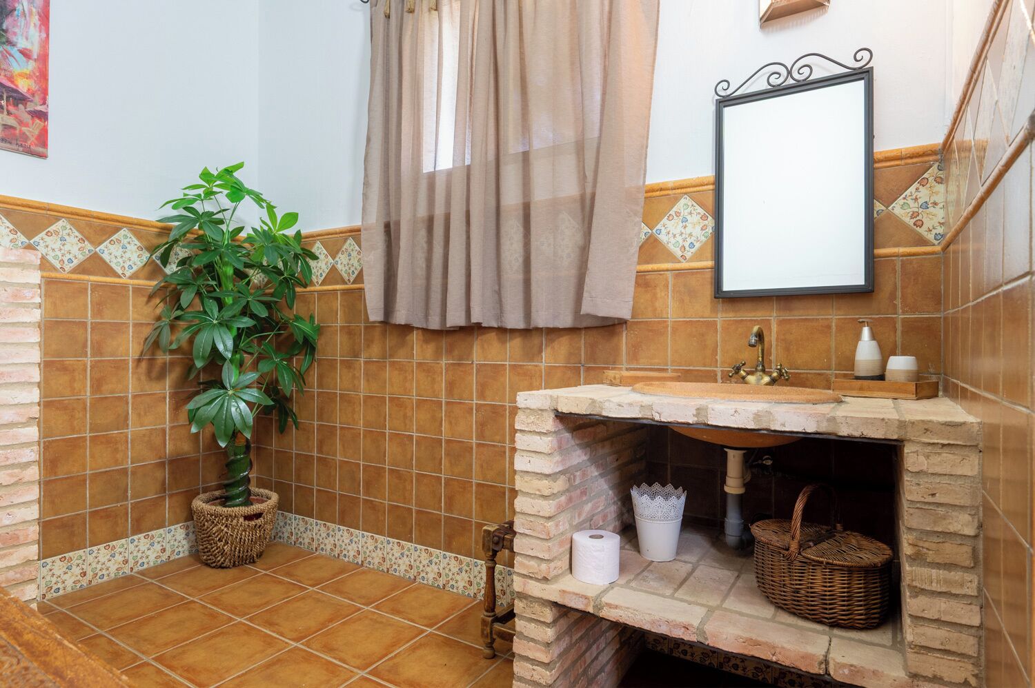 Un baño con piso de baldosas, un lavamanos con base de ladrillo, una planta en maceta, un espejo colgante y una cortina de ducha.