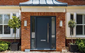 A modern building with a dark entrance door, brick walls, and two potted plants on either side. - Large 6BR House near Goodwood, Garden & Parking (Chichester)