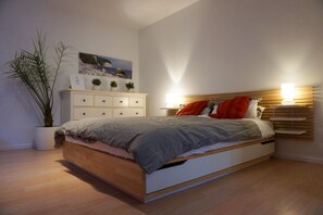 Ein modernes Schlafzimmer mit einem hölzernen Bett, einer weißen Kommode und einer Topfpflanze.