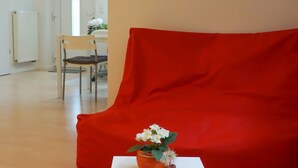 Een kamer met een rode bank, een witte tafel met een bloevas, en twee ingelijste foto's aan de muur.