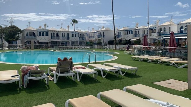 Ett poolområde med solstolar, en swimmingpool och byggnader i bakgrunden.