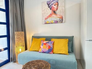 Une pièce avec un lit, un coussin coloré, une table d’appoint en osier et un tableau accroché au mur.