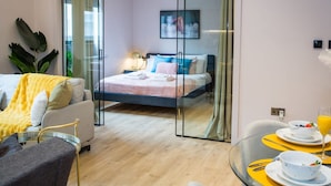 A modern hotel room with a glass partition, a bed with blue bedding, a dining area with yellow placemats, and a plant. - Chic Manchester Chinatown Stay Shops Nightlife (Manchester)