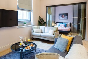 Uma sala de estar moderna com um sofá, uma mesa de centro com itens de café da manhã, uma televisão e uma porta de vidro deslizante que dá para um quarto.
