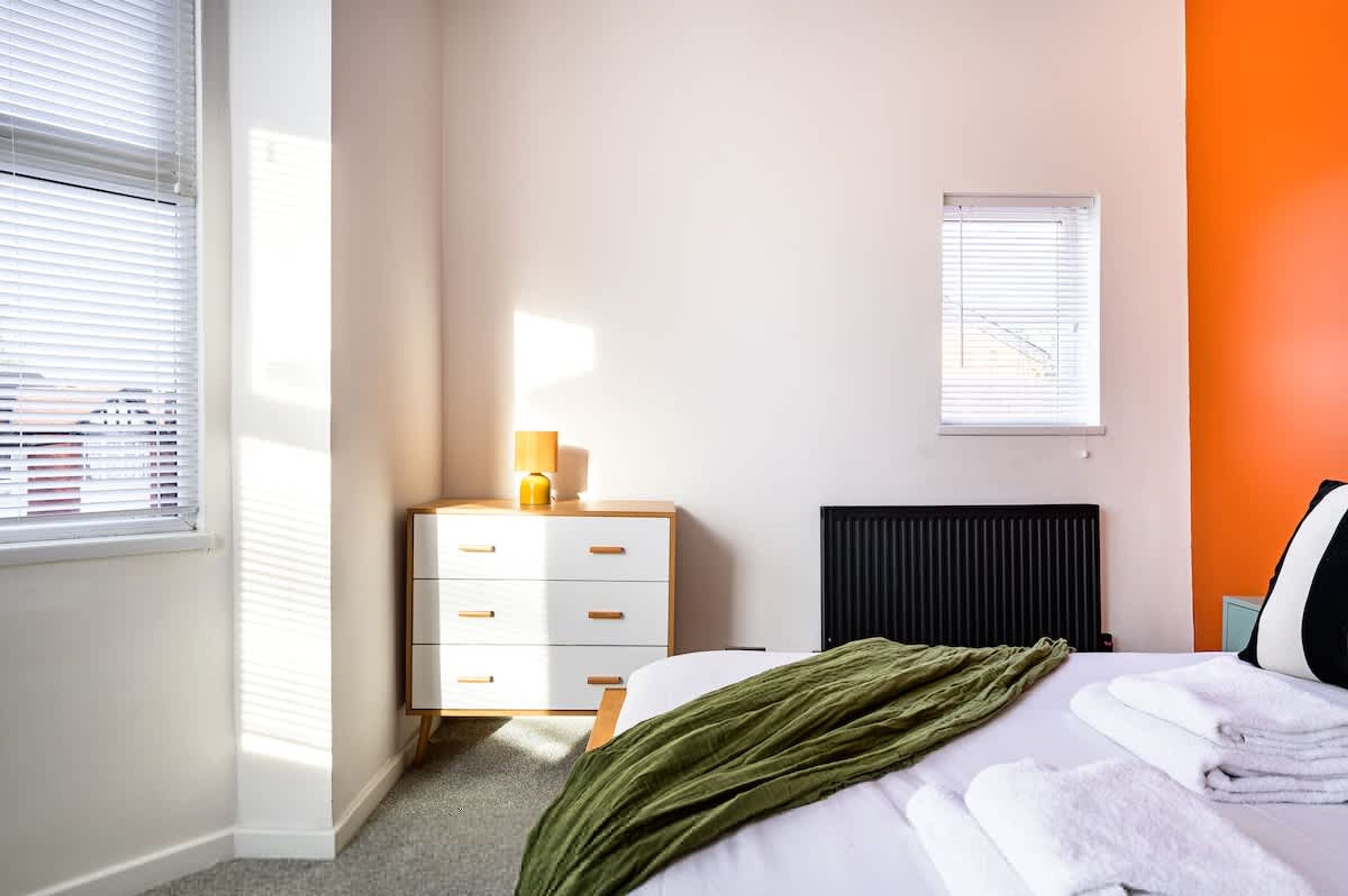 Un dormitorio con cama, mesita de noche con lámpara, radiador y ventana con persianas.