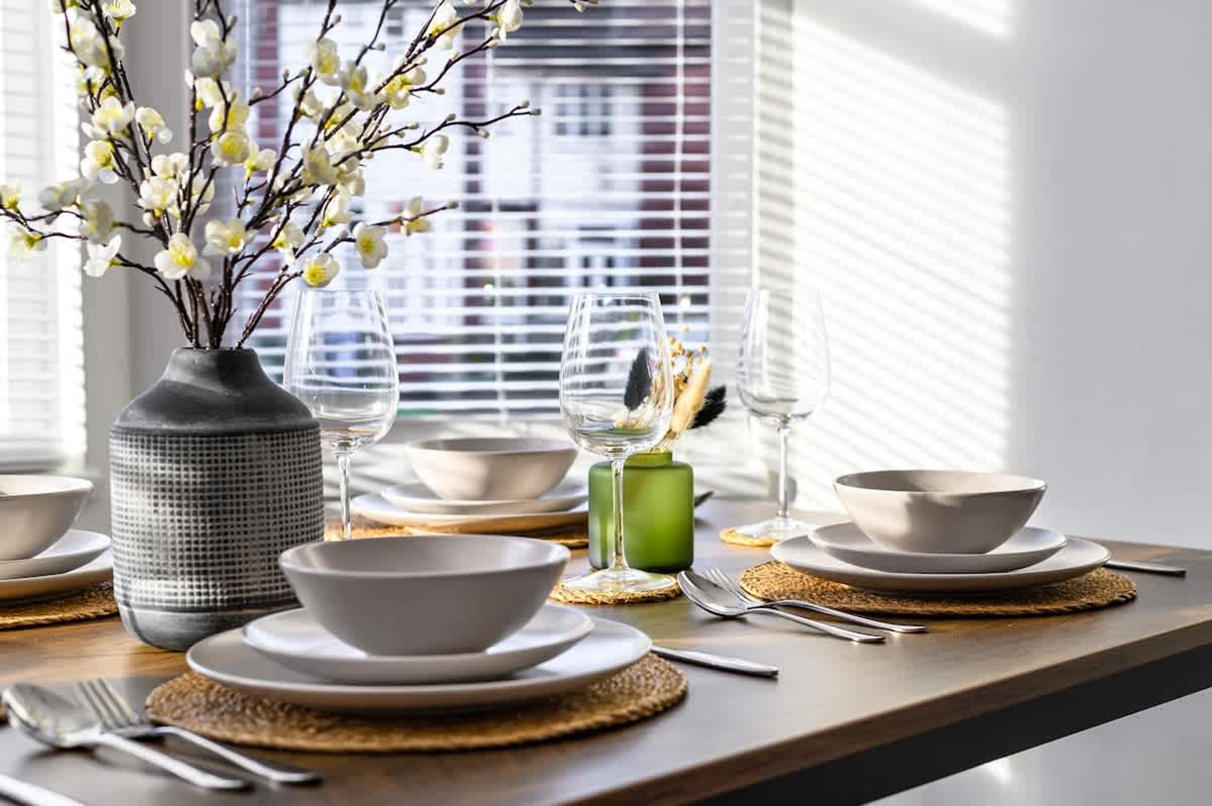 A dining table set with white plates, bowls, and cutlery, a vase with white flowers, and a green candle.