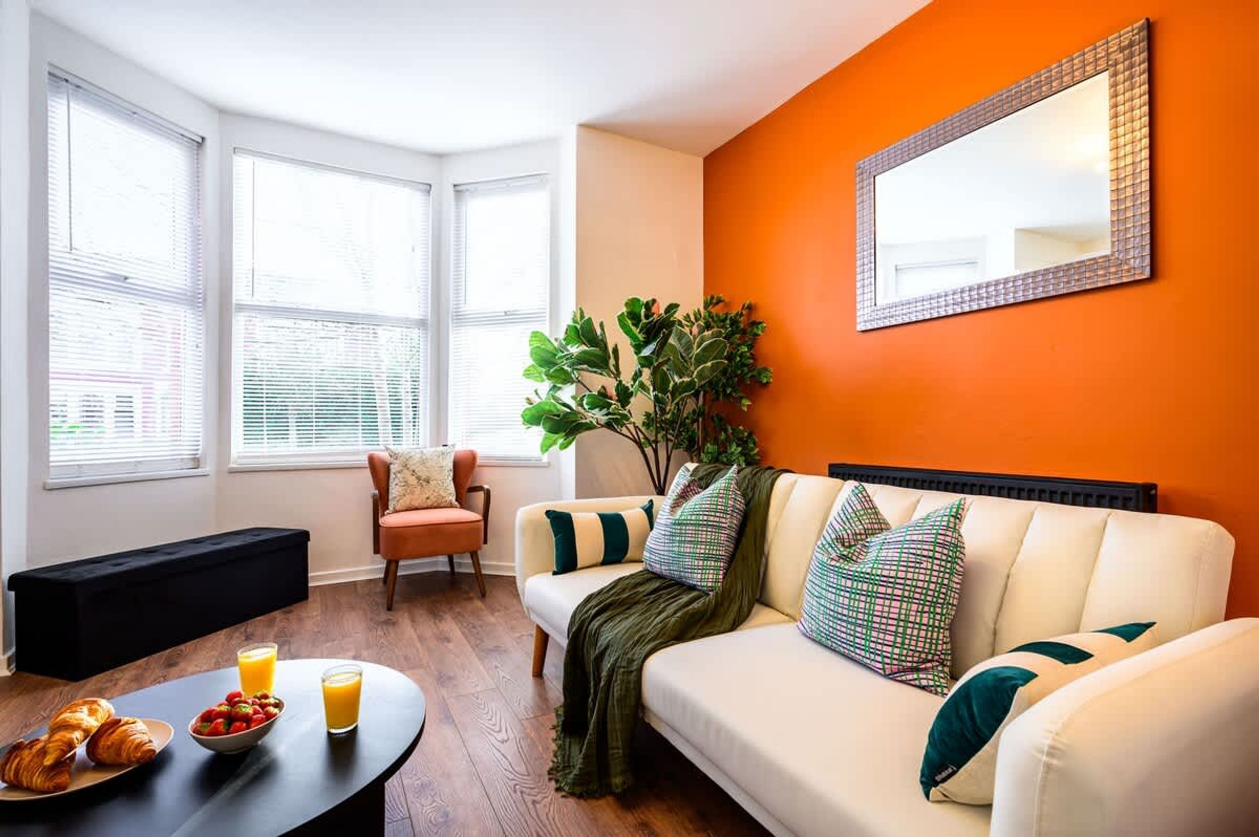A living room with a sofa, a bench, a coffee table with croissants and juice, and a large mirror on an orange wall.