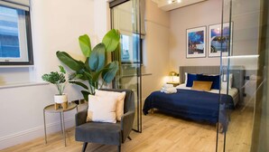 Ein modernes Hotelzimmer mit Glastrennwand, einem Bett mit blauer Bettwäsche, einem grauen Sessel und einer großen Pflanze im Topf.