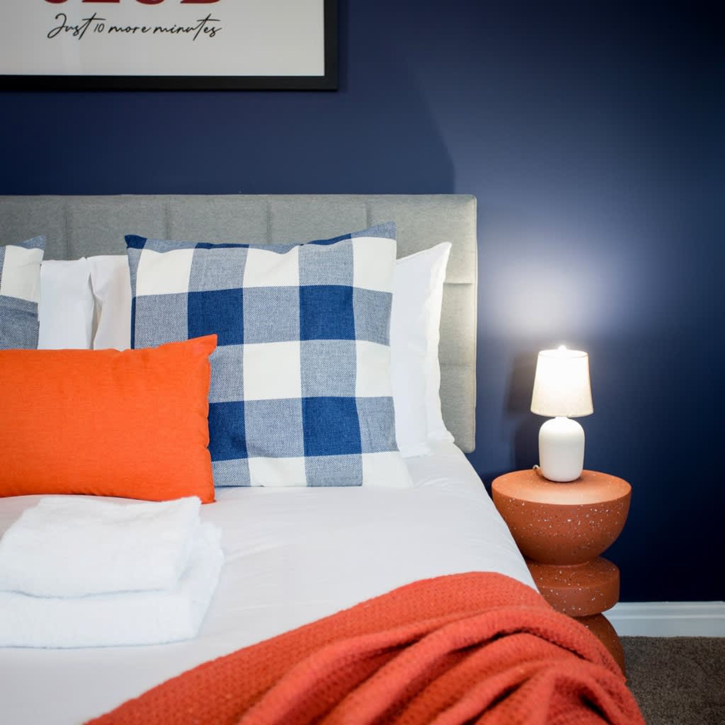 Een opgemaakt bed met een geruit kussen, een wit kussen en een oranje kussen. Er staat een witte lamp op een terracottovoet naast het bed.