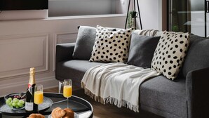 Olohuone, jossa on harmaa sohva, kuviollisia koristetyynyjä, peitto, pieni pöytä, jolla on croissantteja, samppanjapullo ja lasit täynnä appelsiinimehua.