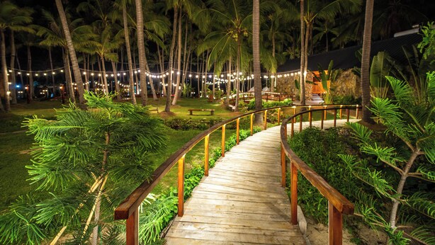 A wooden pathway with railings, surrounded by palm trees and greenery, leading to a building with a thatched roof. - Nirvana Beach Villages (Mui Ne)