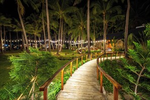 A wooden pathway with railings, surrounded by palm trees and greenery, leading to a building with a thatched roof. - Nirvana Beach Villages (Mui Ne)