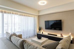 Ein modernes Wohnzimmer mit Sofa, Couchtisch, Fernseher und einem großen Fenster mit transparenten Vorhängen.