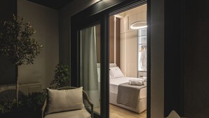 Une chambre d’hôtel moderne dotée d’une porte vitrée donnant sur un balcon, d’un lit avec une tête de lit blanche et d’un petit coin salon avec une table ronde et deux chaises.