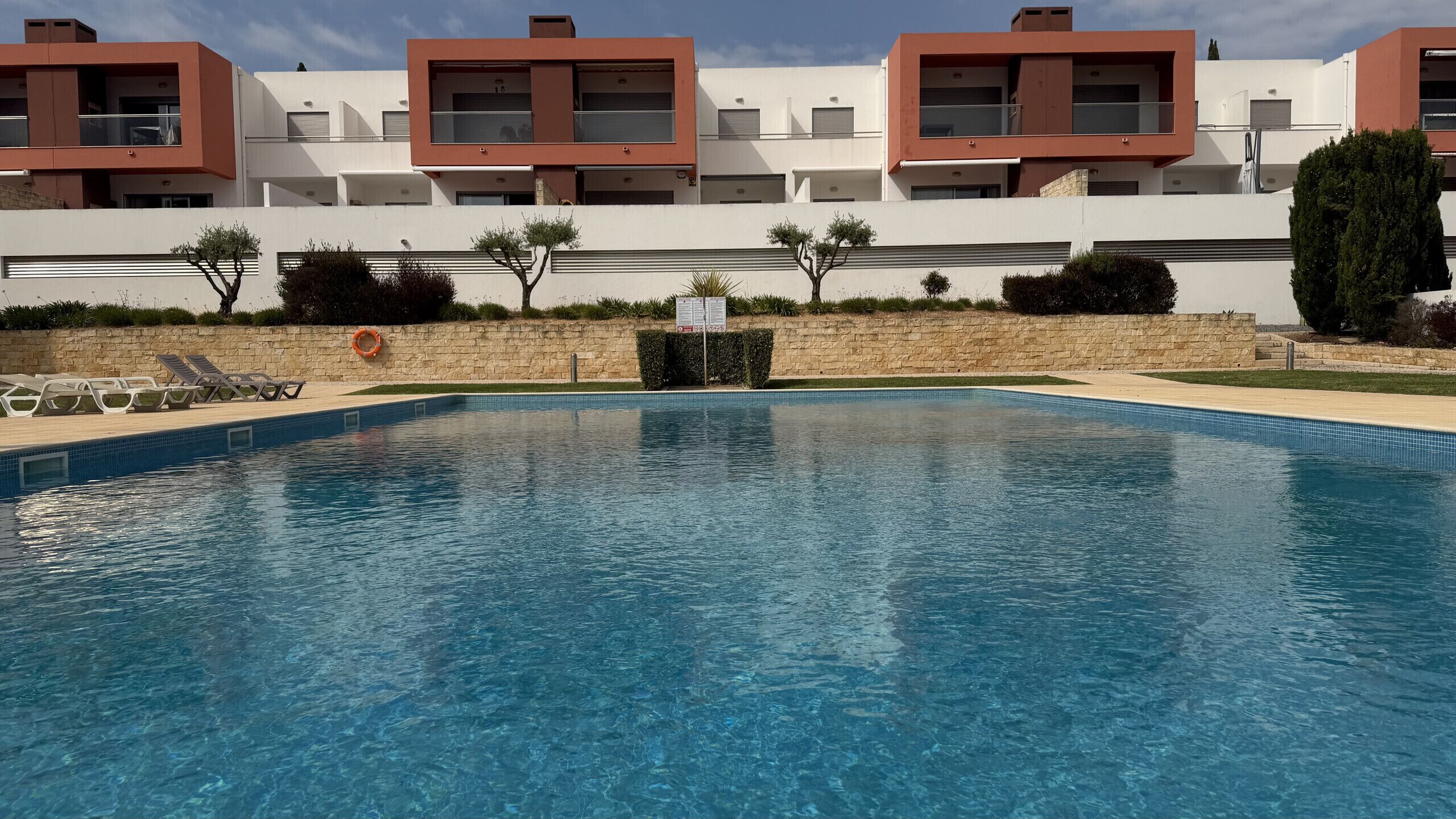 A swimming pool with clear blue water, surrounded by a stone wall and a row of white buildings with brown accents.