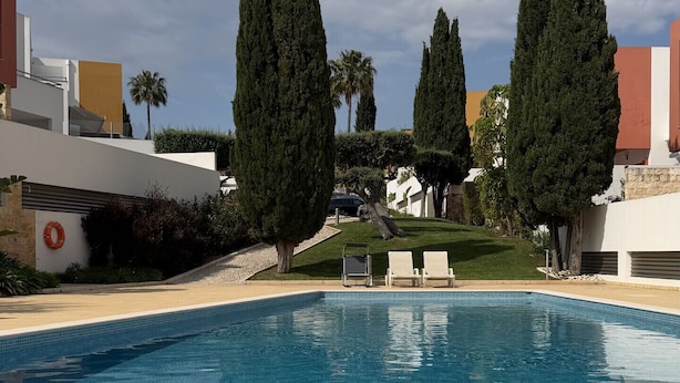 Ein klarer, blauer Swimmingpool mit Liegestühlen, hoch aufragenden Zypressen und modernen Gebäuden im Hintergrund.