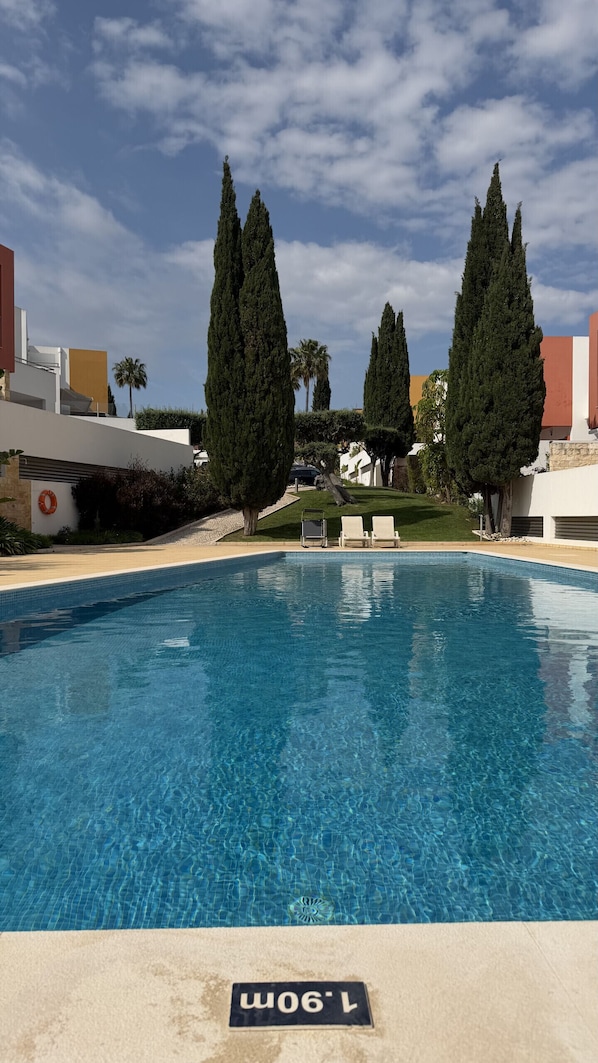 A clear blue swimming pool with lounge chairs, tall cypress trees, and modern buildings in the background. - 2 bedroom apartment, Vitismar, Vale Parra (Albufeira)