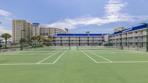 Een tennisbaan met net, omringd door een hek, grenzend aan een hotel met meerdere verdiepingen en balkons.