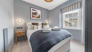 Een slaapkamer met een bed, nachtkastje, lamp, raam en kunstwerk.