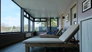 Een kamer met grote ramen, een persoon die op een lounge fauteuil ligt, en uitzicht op bomen en gebouwen.