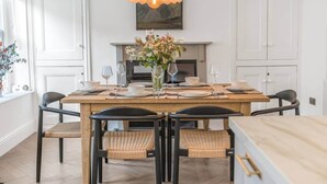 Een eetkamer met een houten tafel gedekt voor vier personen, zwarte stoelen met gevlochten zittingen, een vaas met bloemen en een hanglamp.