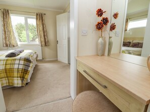 A bedroom with a bed, a bedside table, a mirror, and a window with curtains. - Honeysuckle Lodge (Abergele)