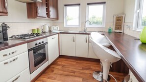 Une cuisine équipée d’une cuisinière à gaz, d’un four, de meubles blancs, d’un plan de travail en bois, d’un évier et de deux fenêtres.