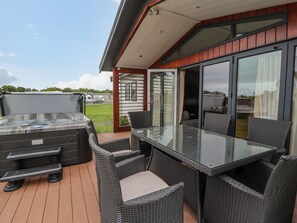 A patio with a glass table and wicker chairs, a hot tub, and a sliding glass door leading to a covered area. - Honeysuckle Lodge (Abergele)