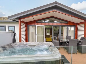 A modern outdoor hot tub area with a covered patio, sliding glass doors, and outdoor furniture. - Honeysuckle Lodge (Abergele)