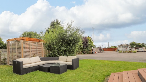 Un espace extérieur verdoyant avec un canapé en osier noir aux lignes courbes, une terrasse en bois et un petit bâtiment entouré d’une clôture blanche.