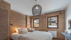 A cozy bedroom with a large bed, wooden walls, two windows, a hanging light fixture, and a radiator. - Renovated and bright apartment - 6 guests (Megève)