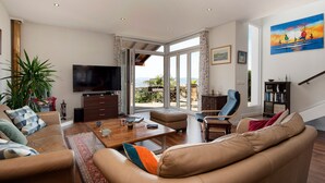A spacious living room with a large TV, comfortable seating, a coffee table, and a view through the glass doors.