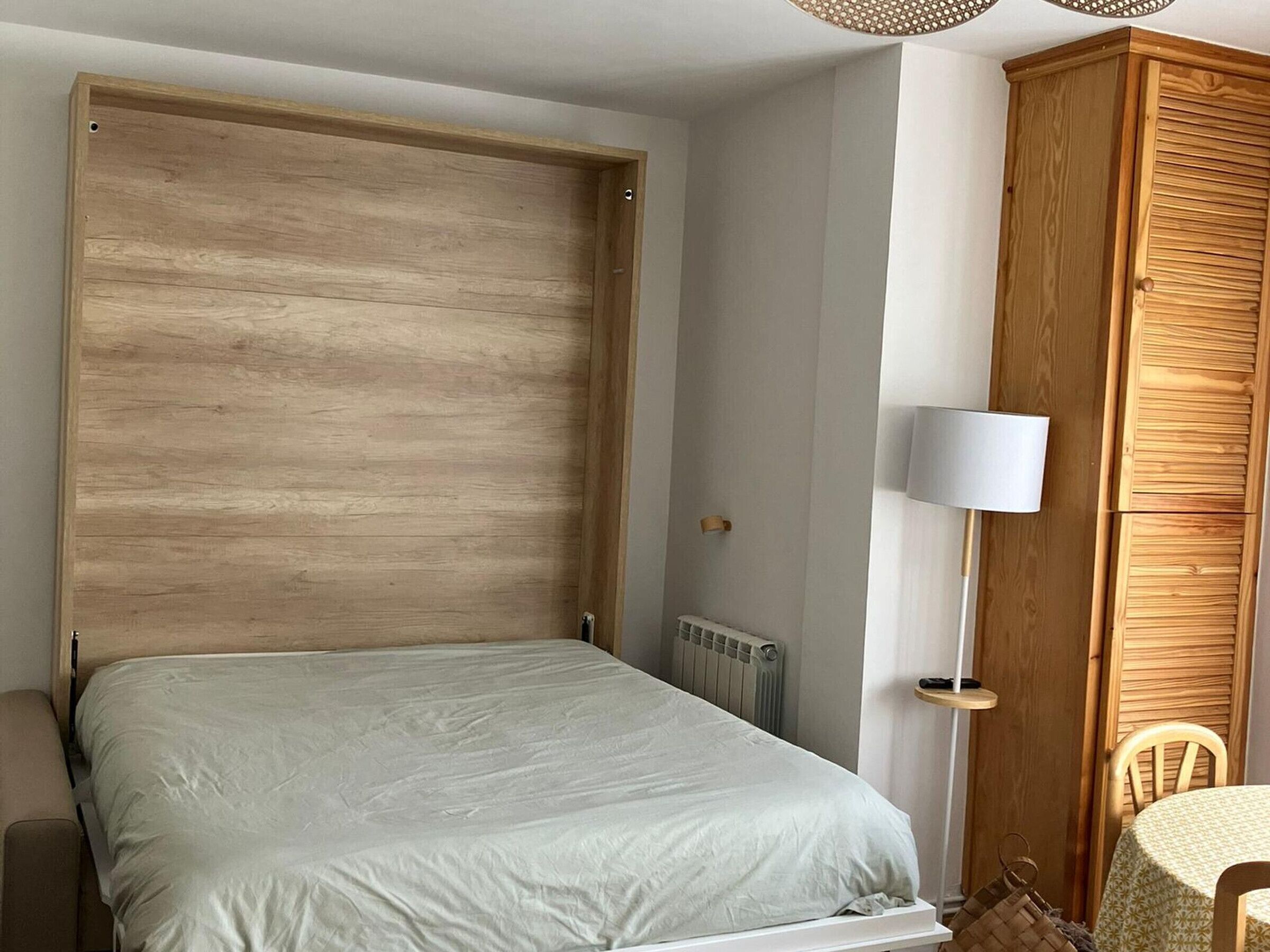 A bedroom with a wooden headboard, a bed with a white sheet, a bedside table with a lamp, and a wooden wardrobe.